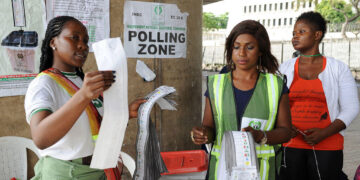 Women election officials stand at an INEC polling zone in Nigeria, handling printed result sheets beside a ballot box during voting