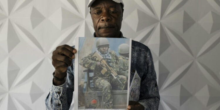 A man wearing a white cap holds up a printed photograph of a soldier in military uniform and body armour, standing against a patterned grey wall.