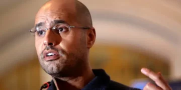 A bald man wearing glasses speaking at a podium, gesturing with his hand while addressing an audience