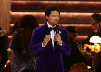 South African comedian Trevor Noah speaking on stage while hosting the 68th Annual Grammy Awards in Los Angeles, wearing a purple velvet tuxedo and holding a microphone