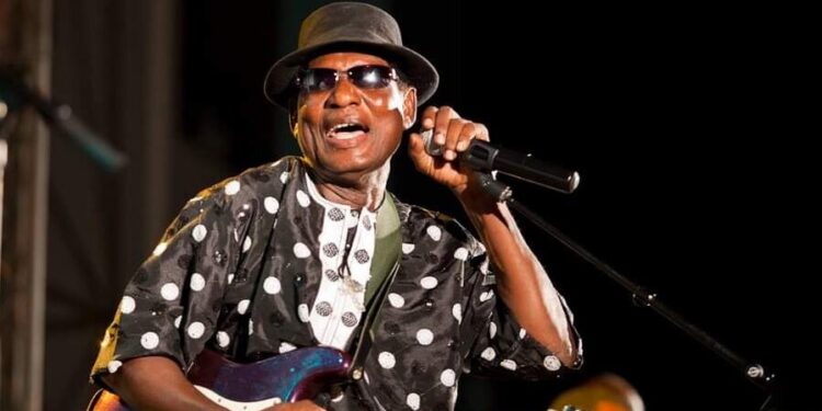 Ebo Taylor performing live on stage with a purple electric guitar and microphone under warm concert lighting