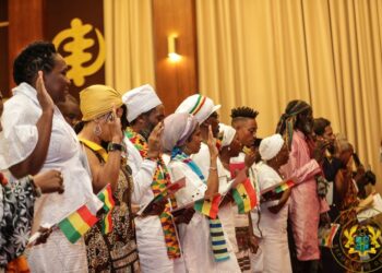 Black members of the African diaspora stand in a line taking the Ghanaian citizenship oath, dressed in white and traditional cloth while holding small Ghana flags in a ceremonial hall in Accra