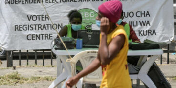 A woman walks past an Independent Electoral and Boundaries Commission voter registration desk in Kenya, where officials sit behind a table with registration equipment and a large IEBC banner in the background.