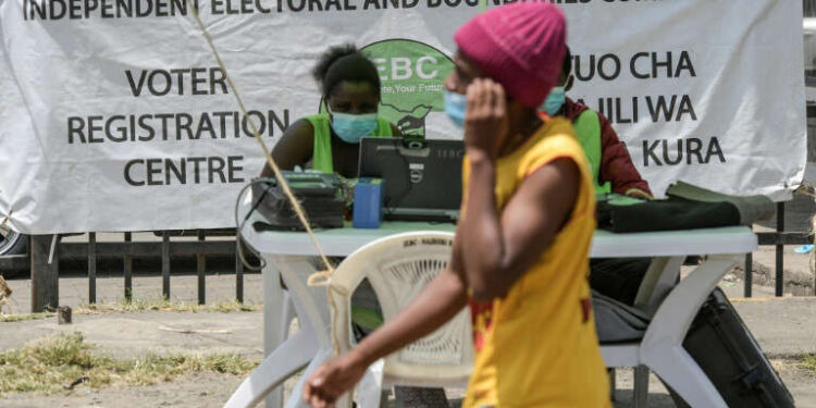 A woman walks past an Independent Electoral and Boundaries Commission voter registration desk in Kenya, where officials sit behind a table with registration equipment and a large IEBC banner in the background.
