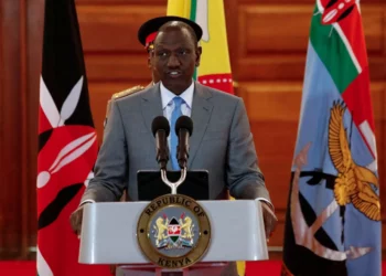 A government official speaking at a podium bearing the Republic of Kenya emblem, flanked by Kenyan national flags inside a wooden-paneled hall