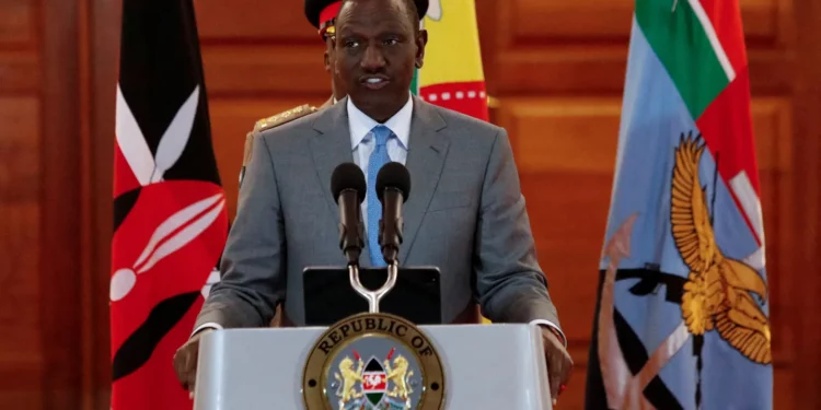 A government official speaking at a podium bearing the Republic of Kenya emblem, flanked by Kenyan national flags inside a wooden-paneled hall