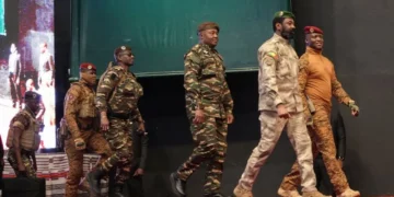 A group of uniformed military officers from the Sahel walking in single file across a red-carpeted stage at a formal public event