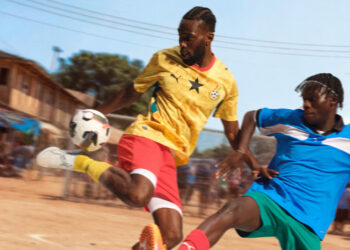 Ghana player in gold Away kit challenges for ball during fast-paced street football match in African urban setting