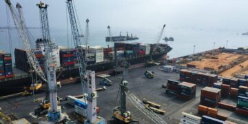Container ships docked at a busy port terminal with cranes loading cargo and stacked shipping containers along the quay