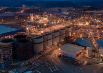 A large-scale gold processing plant in Ghana at dusk, with illuminated industrial facilities, storage tanks and mining infrastructure