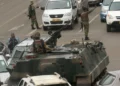 Armed soldiers patrol a city street with an armoured vehicle, stopping traffic during a military security operation in an urban African setting.