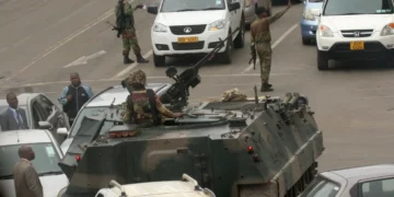 Armed soldiers patrol a city street with an armoured vehicle, stopping traffic during a military security operation in an urban African setting.