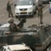 Armed soldiers patrol a city street with an armoured vehicle, stopping traffic during a military security operation in an urban African setting.
