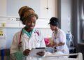 African healthcare worker using a tablet in a hospital ward while a colleague attends to a patient in the background