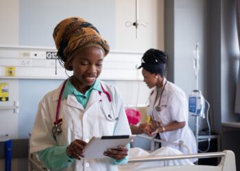 African healthcare worker using a tablet in a hospital ward while a colleague attends to a patient in the background