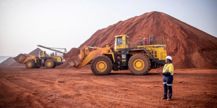 Heavy machinery loads bauxite at a mining site in Guinea, with large red ore stockpiles in the background