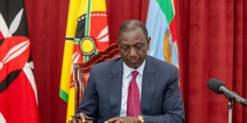 Kenyan President William Ruto seated at a desk signing documents, with Kenyan flags behind him