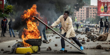 A protester pushes burning tyres onto a road barricade during demonstrations over rising fuel prices in Nairobi, as smoke billows across a street scattered with rocks and debris