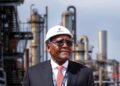 Aliko Dangote wearing a white safety helmet stands in front of refinery infrastructure at an industrial energy facility