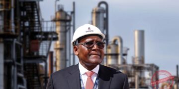 Aliko Dangote wearing a white safety helmet stands in front of refinery infrastructure at an industrial energy facility