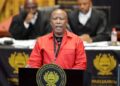 Julius Malema wearing a red EFF uniform while addressing South Africa’s parliament from a podium