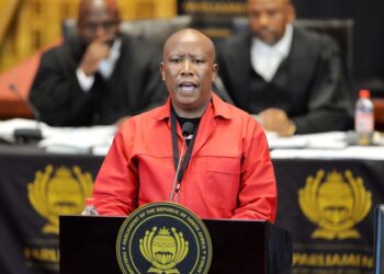 Julius Malema wearing a red EFF uniform while addressing South Africa’s parliament from a podium