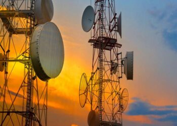 Telecommunications towers with satellite dishes and wireless antennas silhouetted against a sunset sky, representing broadband and mobile network infrastructure expansion in West Africa