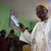 Cameroonian opposition figure Issa Tchiroma Bakary holds up a ballot paper after voting at a polling station during the 2025 presidential election, as journalists and observers watch