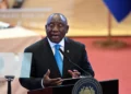 South African President Cyril Ramaphosa speaks at a podium during a public address, wearing a dark suit and blue tie, with the presidential seal visible and a blurred official backdrop behind him