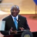 South African President Cyril Ramaphosa speaks at a podium during a public address, wearing a dark suit and blue tie, with the presidential seal visible and a blurred official backdrop behind him