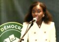 Mariama Djibrine speaks at a podium into a microphone while wearing a white double-breasted blazer, with a green backdrop displaying the logo of the Alliance des Démocrates du Sahel