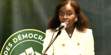 Mariama Djibrine speaks at a podium into a microphone while wearing a white double-breasted blazer, with a green backdrop displaying the logo of the Alliance des Démocrates du Sahel