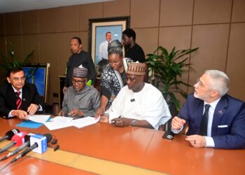 Officials from Africa Finance Corporation and Dangote Group representatives sit around a conference table during a document-signing ceremony, with media microphones in the foreground and staff observing in the background