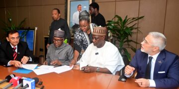 Officials from Africa Finance Corporation and Dangote Group representatives sit around a conference table during a document-signing ceremony, with media microphones in the foreground and staff observing in the background