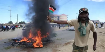 A protester holding a Kenyan flag stands beside burning debris and thick black smoke during demonstrations in Nanyuki, Kenya, against a proposed Ebola isolation facility near Laikipia Airbase