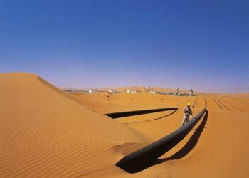 Gas pipeline crossing sand dunes in the Sahara Desert with a worker inspecting infrastructure, representing Algeria's Trans-Saharan Gas Pipeline project linking Nigeria, Niger and Europe