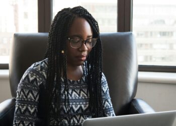 Black woman wearing glasses concentrating on her laptop beside a large office window