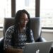 Black woman wearing glasses concentrating on her laptop beside a large office window