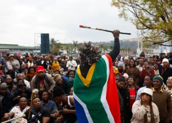 A person draped in the South African flag raises a decorated stick above a large crowd gathered outdoors