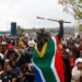 A person draped in the South African flag raises a decorated stick above a large crowd gathered outdoors