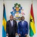 Two government officials stand between the flags of The Bahamas and Ghana beneath the Bahamian coat of arms